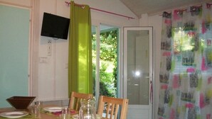 A dining area with a wooden table set for four, chairs, a television mounted on the wall, and a sliding glass door leading to a garden. - ZEcamping 3* familial pataugeoire dog friendly locatif LV BBQ 40m2 (La Souterraine)