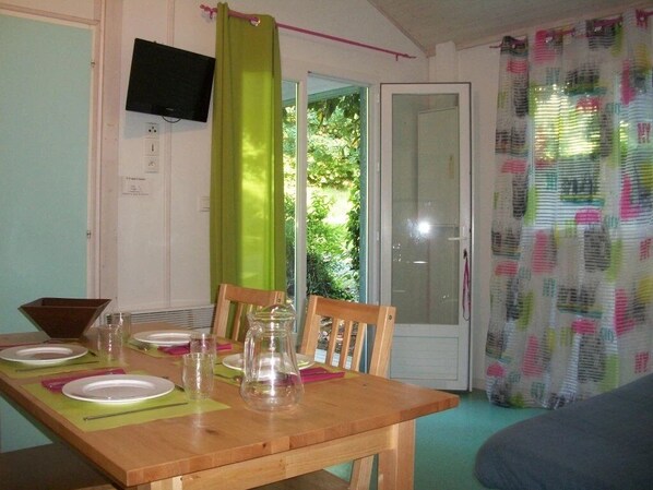 A dining area with a wooden table set for four, chairs, a television mounted on the wall, and a sliding glass door leading to a garden.
