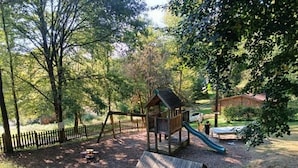 A playground with a slide and a wooden structure, surrounded by trees and a fenced area.