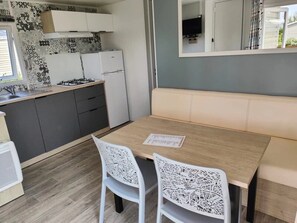 Eine kleine Küche mit Gasherd, Kühlschrank und Spüle. Ein Holztisch mit zwei Stühlen und einer Bank steht bereit.