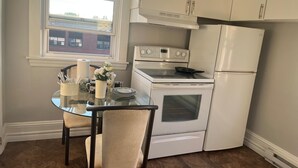 A compact kitchen with a stove, oven, and refrigerator, a small dining table with chairs, and a window with a view of buildings outside.