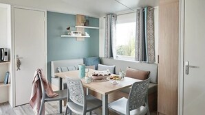 A modern dining area with a wooden table, grey chairs, a bench, and a window with patterned curtains. - Camping 3 étoiles - Piscine  - ccbfecg (Penvénan)