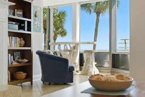 A room with a blue armchair, a wooden bookshelf, a dining table with a white top, and a view of palm trees through large windows. - Palm Beach Club Condo #119 (Pensacola Beach)