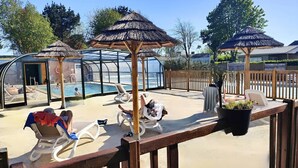 Zona de piscina con sillones de descanso, piscina cubierta con paredes de vidrio y sombrillas de paja.
