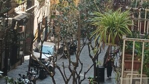 Een smalle straat met gebouwen aan beide kanten, bloempotten en geparkeerde scooters.