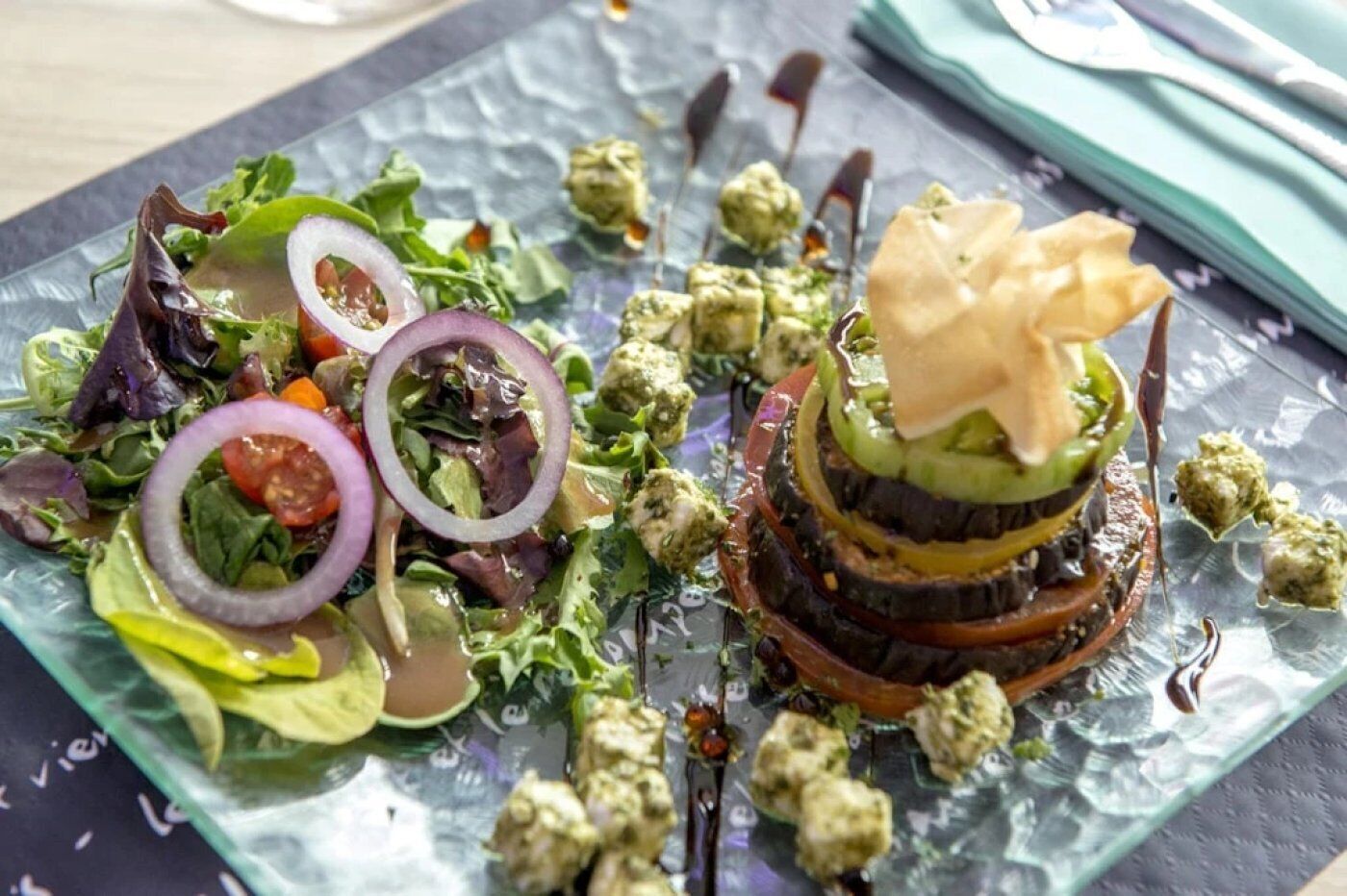 Un piatto di vetro con un'insalata e un piatto a strati, forse un hamburger, con una forchetta e un tovagliolo a lato.