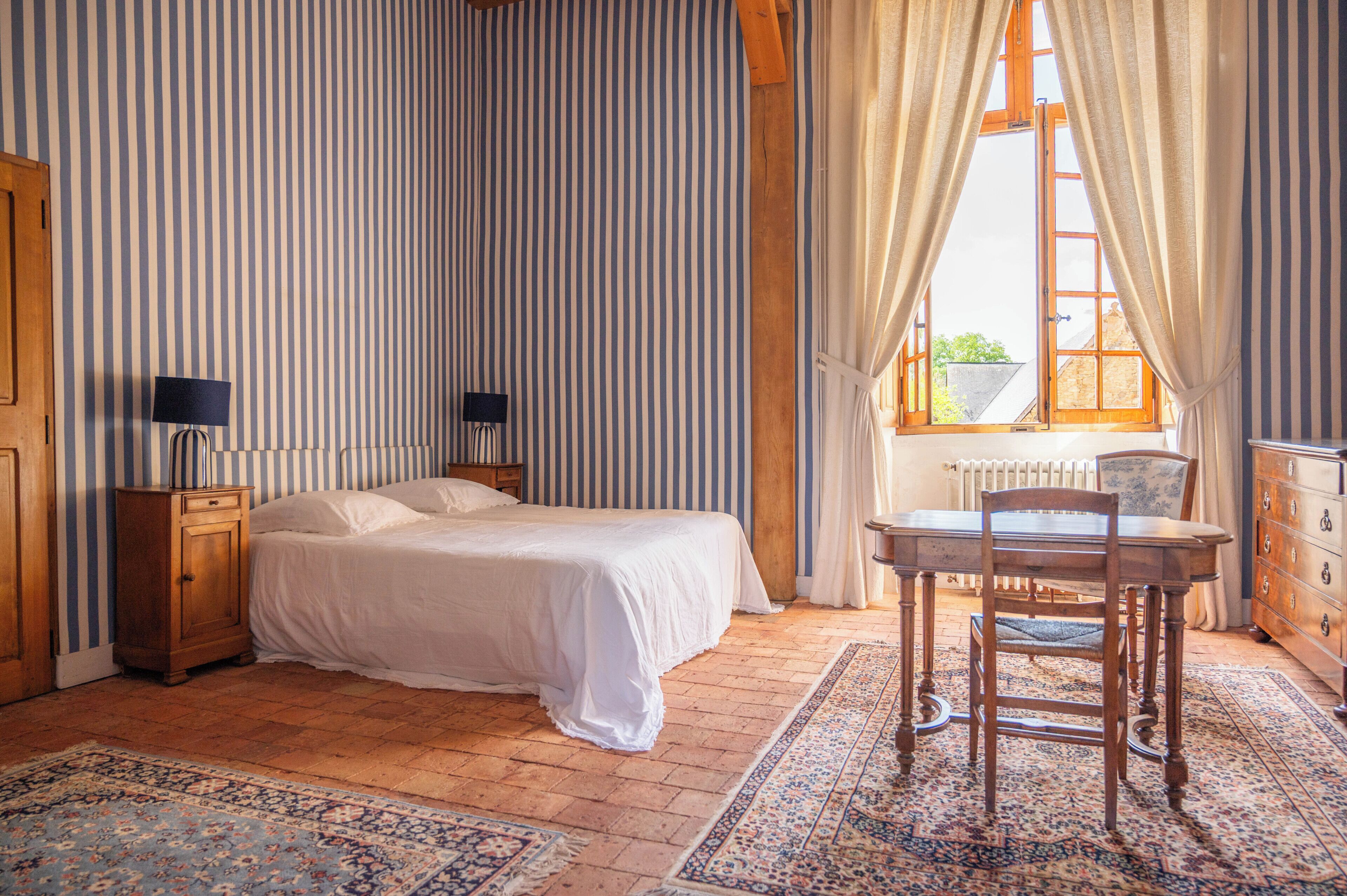 Un dormitorio con cama, una mesita de noche de madera, una mesa con silla, un radiador y una ventana con cortinas.