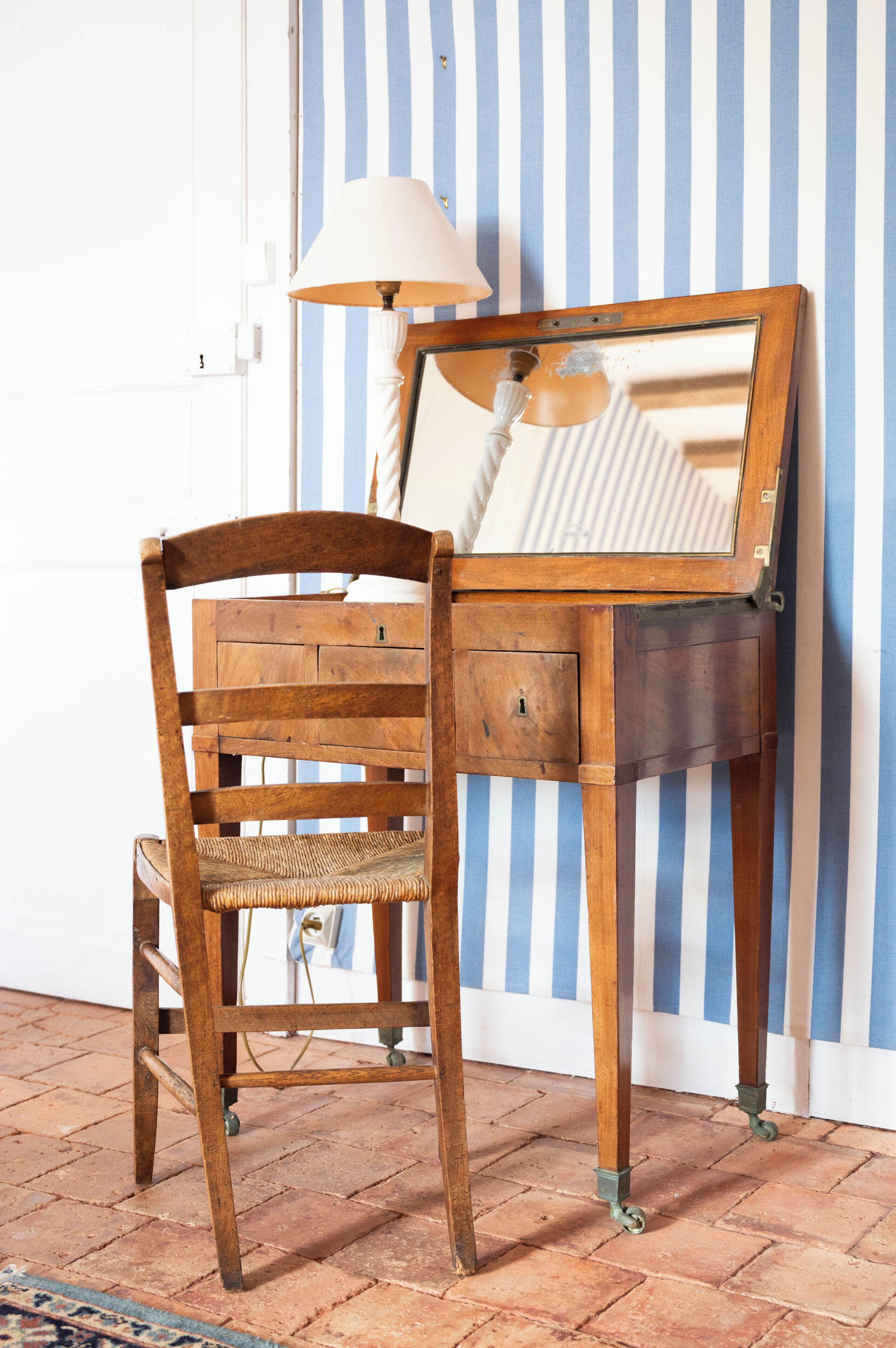 A wooden chair with a woven seat and backrest, a small wooden table with a mirror and lamp, and a striped wall in the background.