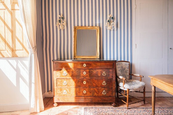 A room with a wooden dresser, a chair, a mirror, and striped wallpaper.