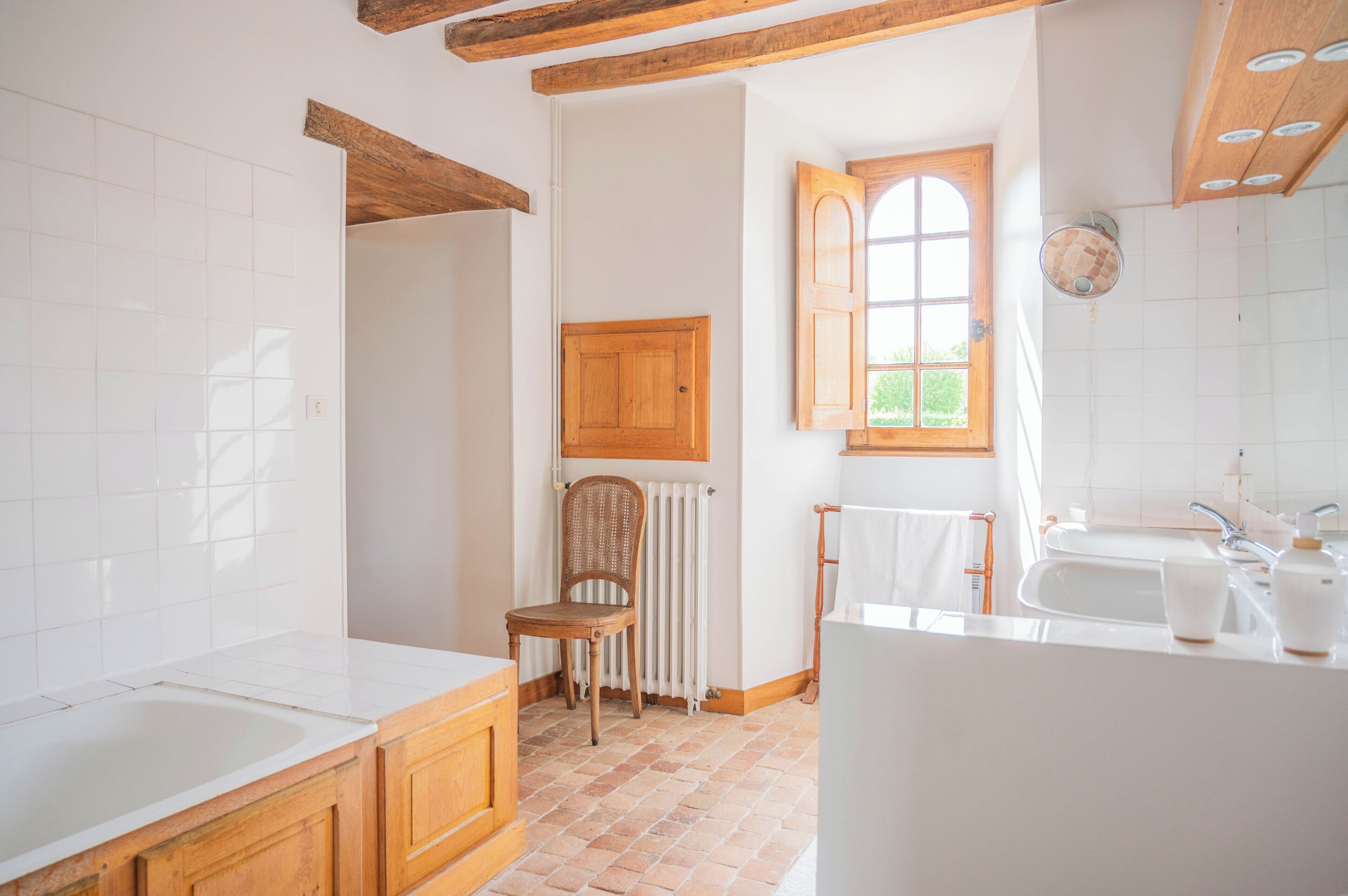 Een badkamer met een houten stoel, een radiator, een raam met open luiken en een witte wastafel met zeepdispenser.