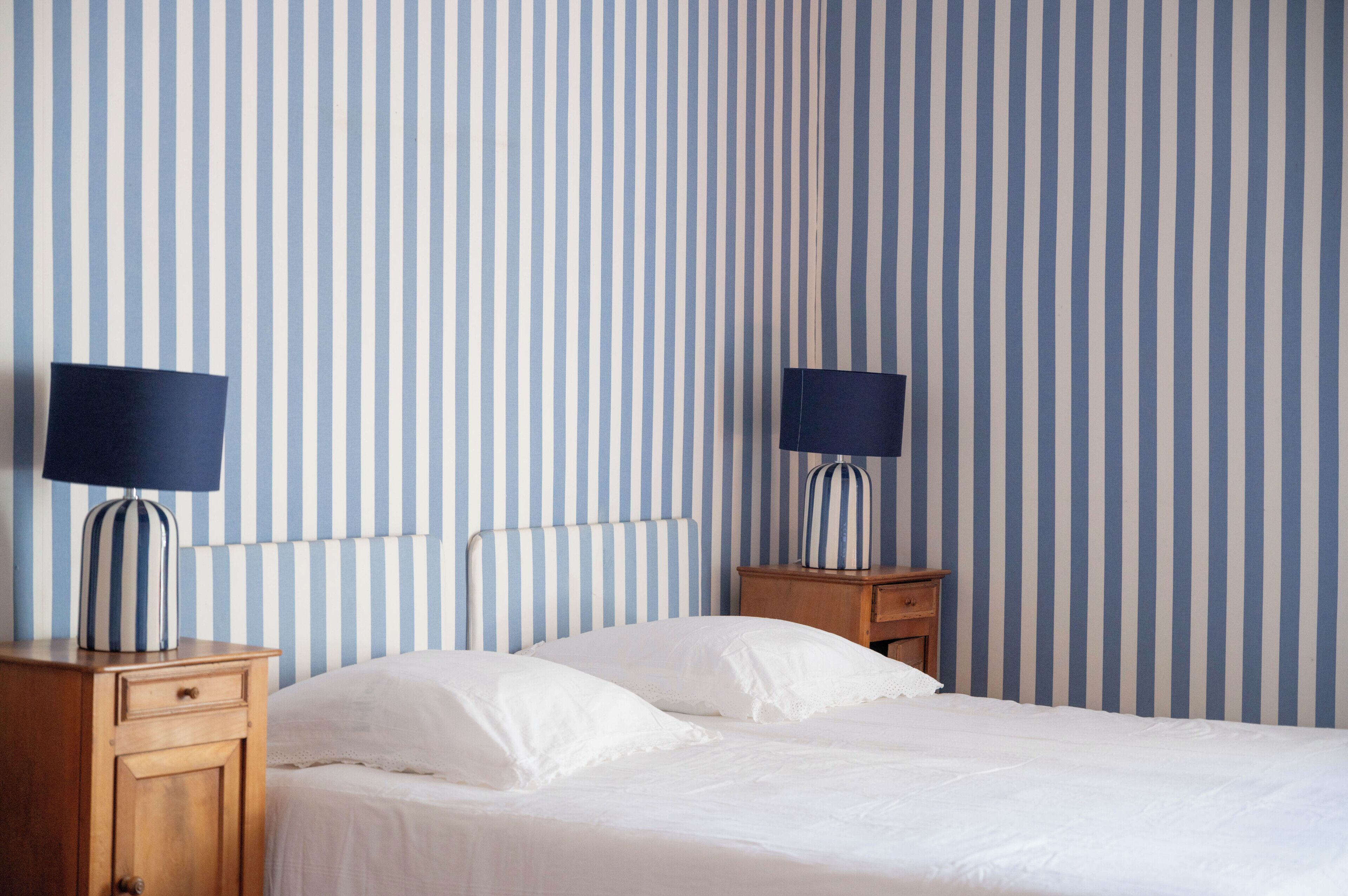 Una habitación de hotel con pared a rayas, una cama con sábanas blancas, dos mesitas de noche con lámparas y un tocador de madera.