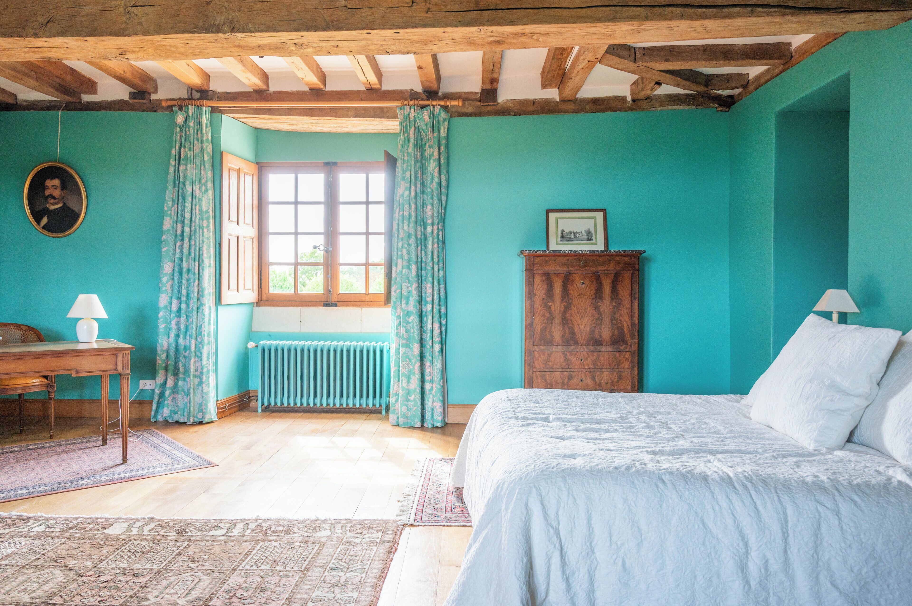 A bedroom with a bed, a wooden cabinet, a desk, a chair, a radiator, and a window with curtains.