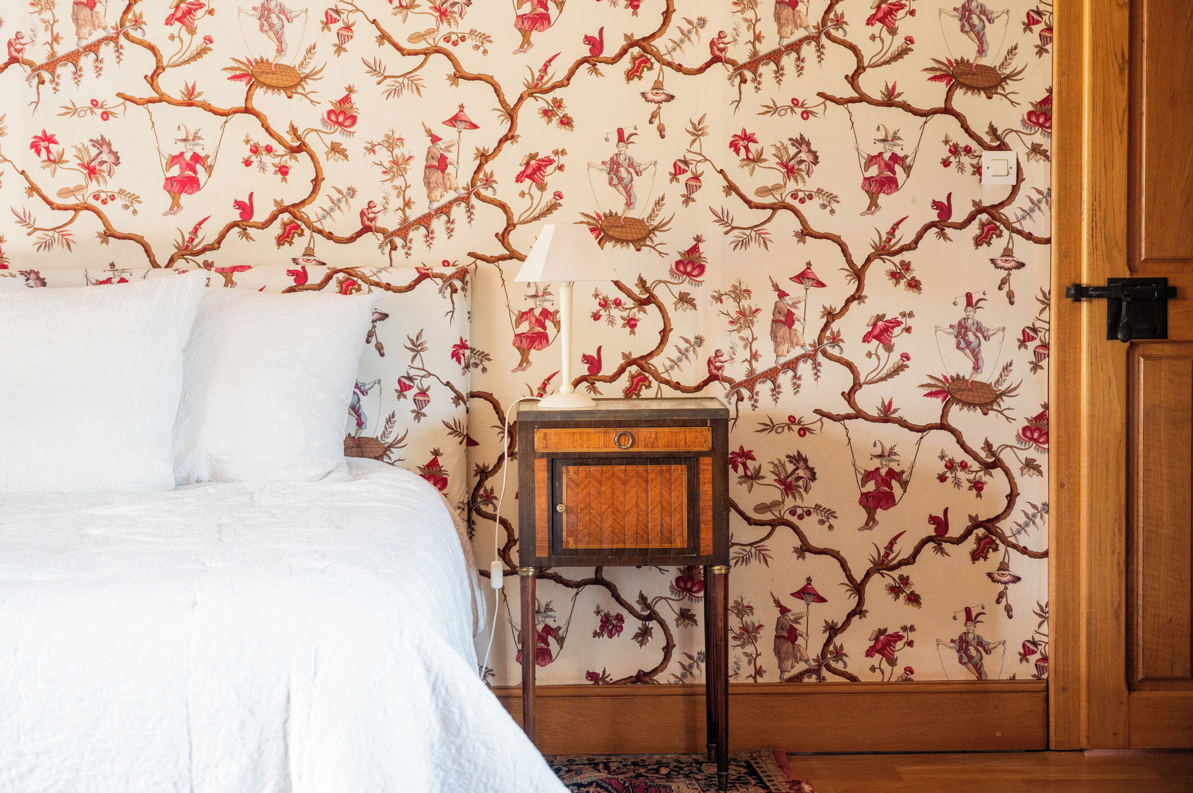 A bedroom with a bed, a nightstand, a lamp, and a floral wallpaper.
