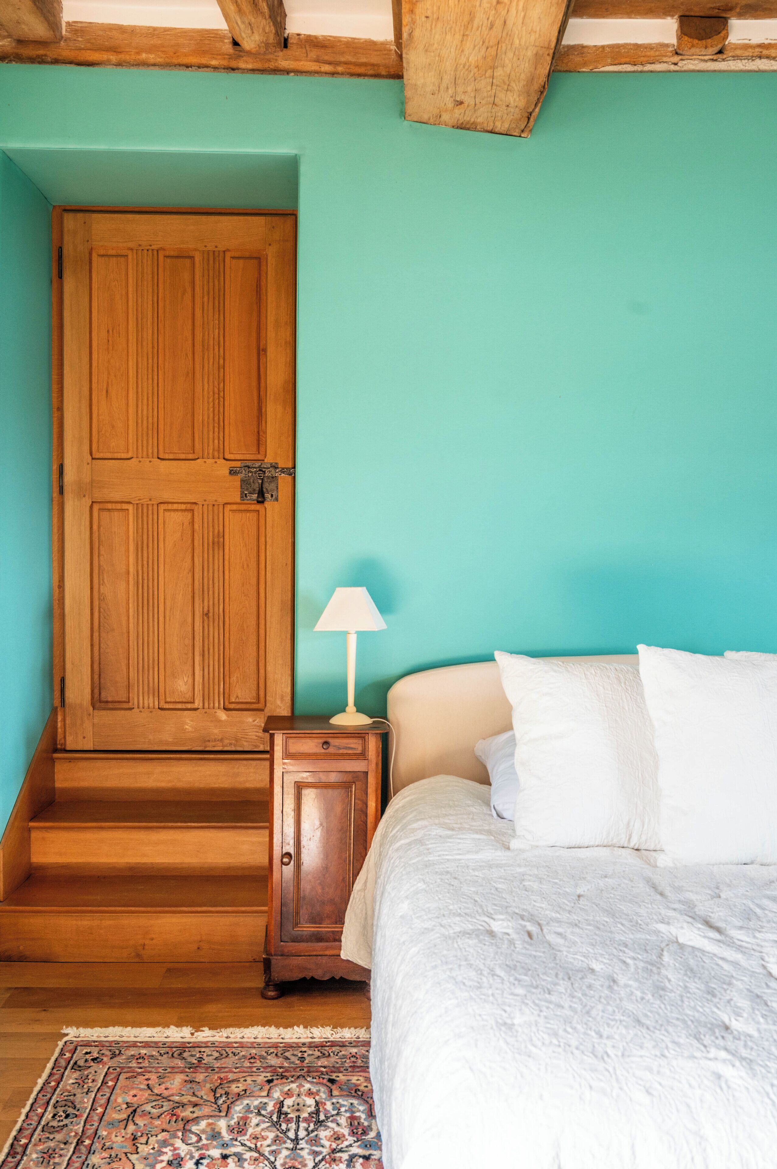 A bedroom with a wooden door, a nightstand with a lamp, a bed with white bedding, and a patterned rug.
