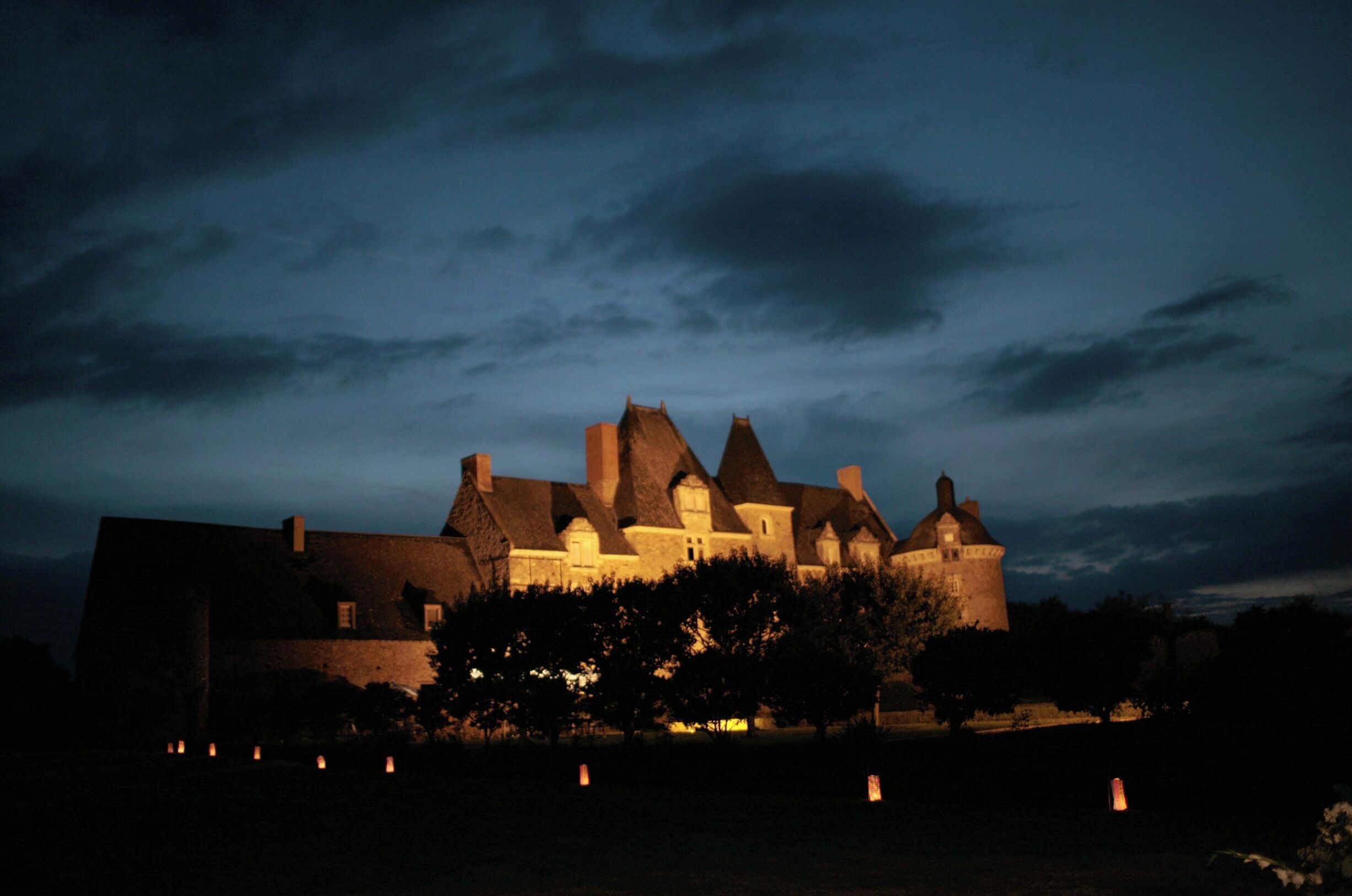 Un grand bâtiment historique doté de nombreuses tours, dont une tour centrale imposante, entouré d’arbres et illuminé par un éclairage d’ambiance.