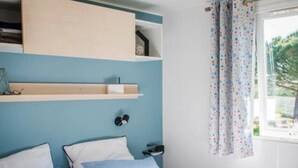 A bedroom with a bed, pillows, a bedside table, a shelf, and a window with curtains. - Camping 3 étoiles - Piscine  - ccbfgbf (Collias)