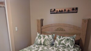 Una cama con cabecera de madera, colcha con estampado de hojas tropicales y un cuadro de palmeras colgado en la pared.