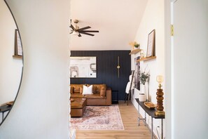 Een gang die uitkomt in een woonruimte met een bruine leren bank, een kamerbreed tapijt, een plafondventilator en een wandplank met decoratieve objecten.