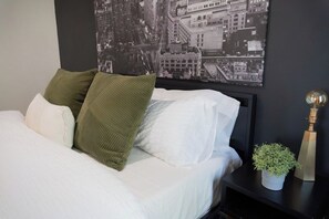 Een net opgemaakt bed met witte beddengoed en twee kussens, een zwart nachtkastje met een kleine plant in pot, en een zwart-wit foto van een stadsgezicht op de muur.