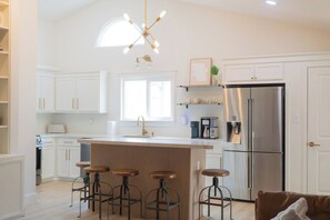 A modern kitchen with white cabinetry, a central island with stools, a stainless steel refrigerator, and a pendant light. - A Cozy Retreat | Near Austin | Pet Friendly (Cedar Park)