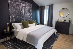 A modern bedroom with a large bed, a cityscape artwork, a dresser, and a mirror. - A Cozy Retreat | Near Austin | Pet Friendly (Cedar Park)