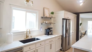 A modern kitchen with white cabinetry, a stainless steel refrigerator, a built-in oven, a sink with a gold faucet, and a window providing natural light. - Cozy 3BR/2BA - Peaceful, Pet-friendly Home (Cedar Park)