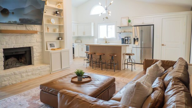 A modern living room with a fireplace, a leather sofa, a coffee table, and a kitchen area with a refrigerator and bar stools. - Cozy 3BR/2BA - Peaceful, Pet-friendly Home (Cedar Park)