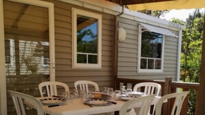 Un espacio comedor al aire libre con techo, una mesa y sillas, piso de madera y un pequeño edificio con ventana y toldo.