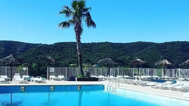 Piscina azul clara con sillones de descanso y una palmera, rodeada por una cerca y un paisaje montañoso.