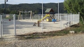Ein umzäunter Spielplatz mit Rutsche und Klettergerüsten, umgeben von Kies und Bäumen.