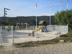 A fenced playground with a slide and climbing structures, surrounded by a gravel area and trees. - Camping 3 étoiles - Piscine  - ccbfgcg (Collias)