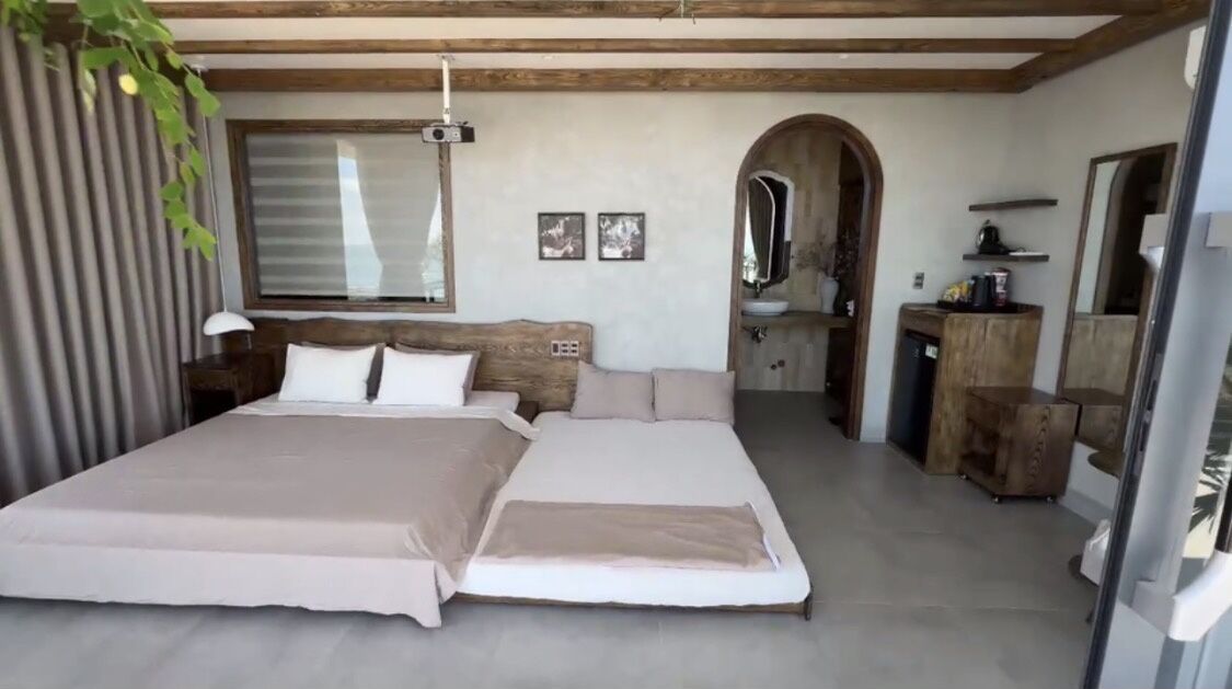 A bedroom with a large bed, a wooden headboard, a bedside table, a mirror, and a small kitchenette area.