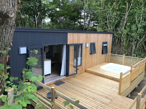 A modern cabin with a wooden deck, a hot tub, and a glass door entrance.