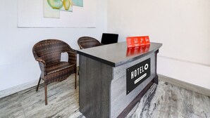 Ein Empfangstresen mit einem Schild, auf dem 'HOTEL' und ein Hinweis 'Company Serviced' stehen, zwei Korbstühle und ein Wandbild.
