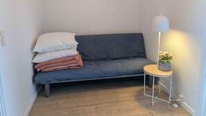 Quarto pequeno e acolhedor com um sofá cinza, uma mesa de cabeceira com abajur e uma pequena planta em vaso.