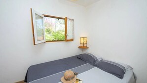 Una cama individual con una colcha a rayas grises y blancas, una mesita de noche con una lámpara, una ventana con una cortina abierta y un sombrero sobre la cama.