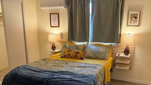 Une chambre à coucher avec un lit, des lampes de chevet, une fenêtre avec des rideaux, un climatiseur fixé au mur et un tableau encadré.