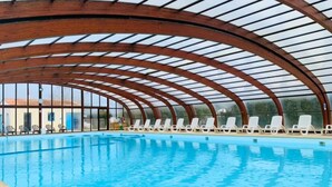 An indoor swimming pool with a curved wooden roof and white lounge chairs along the poolside. - Camping 3 étoiles - Piscine  - ccbffbc (Saint-Denis-d'Oléron)