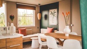 Een moderne woonkamer met een bank, eettafel, stoelen, een hanglamp en een raam met uitzicht op groen.