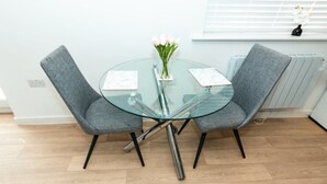 A modern dining area with a glass table, two grey upholstered chairs, a vase with flowers, and a window with blinds. - Nay Apartment Two-Bedroom Appt in Coventry (Longford)