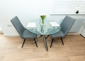 A modern dining area with a glass table, two grey upholstered chairs, a vase with flowers, and a window with blinds. - Nay Apartment Two-Bedroom Appt in Coventry (Longford)
