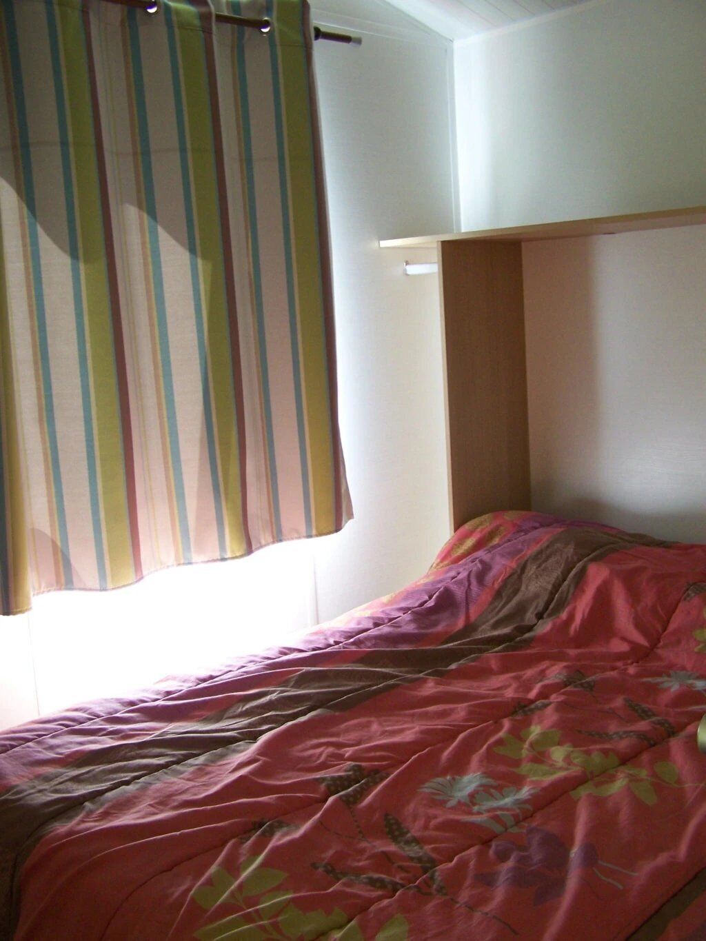 Un lit simple avec une couvre-lit à motifs floraux, un rideau à rayures et un mur uni agrémenté d’une tête de lit en bois.