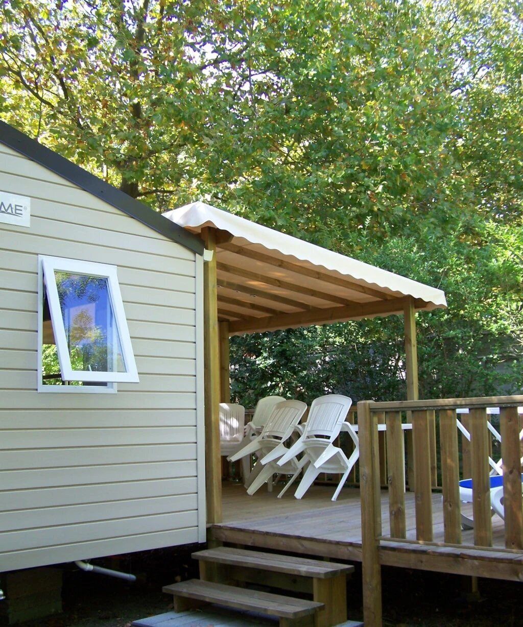 A covered outdoor area with a wooden deck, white plastic chairs, and a small building with a window.
