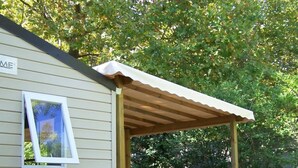 A covered outdoor area with a wooden deck, white plastic chairs, and a small building with a window. - Camping 3 étoiles - Piscine  - ccbffec (Arces)