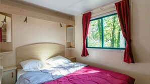 A single bed with a headboard, a window with red curtains, and a shelf unit with a mirror. - Camping 3 étoiles - Piscine  - ccbfg0g (Chauffour-sur-Vell)