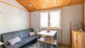 Une petite pièce avec un plafond en bois, un canapé gris, une table en métal avec des chaises, une fenêtre et un meuble en bois.