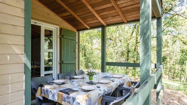 Un espace extérieur abrité, aménagé pour les repas, avec une table dressée pour quatre personnes, entourée de chaises, et offrant une vue sur une zone boisée.
