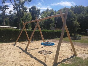 Eine Schaukelanlage in einer sandigen Fläche mit Bäumen im Hintergrund.