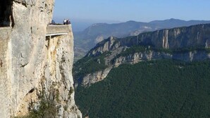 A cliffside with a narrow ledge, overlooking a valley with dense forest and distant mountains.