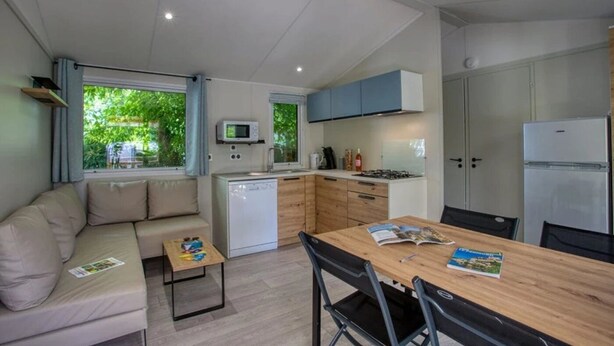 A compact living space with a kitchenette, a sofa, a dining table, and a view of greenery outside. - Camping 3 étoiles - Piscine  - ccbfgee (Groléjac)